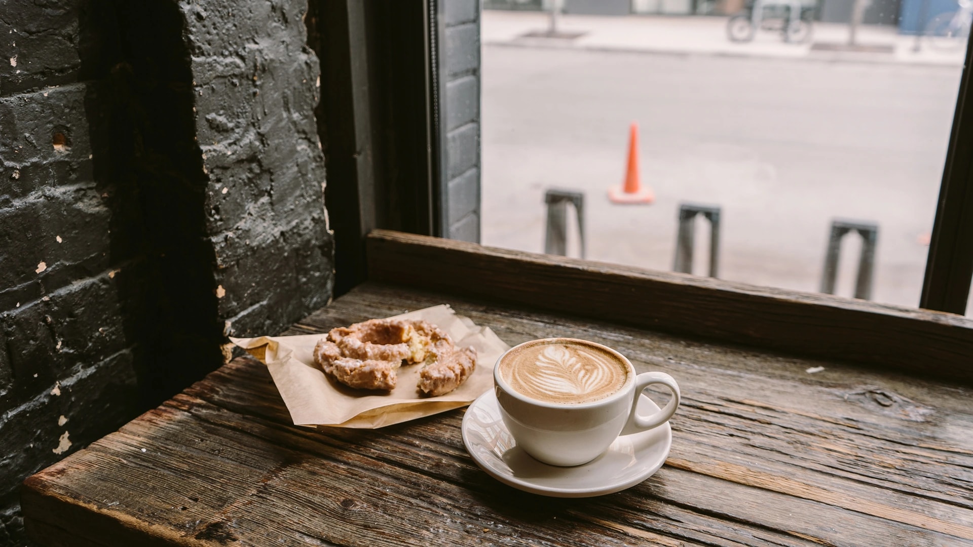 En kopp cappuccino med lattekunst på et rustikt trebord ved et kafévindu, med fersk bakst i bakgrunnen.